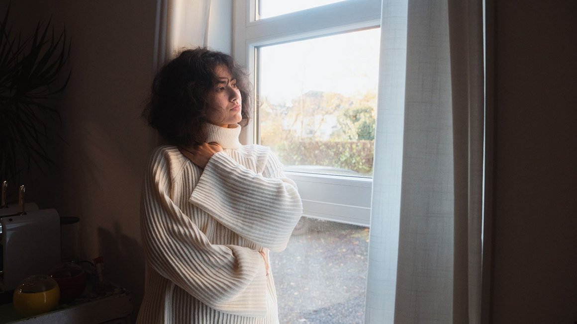 Une femme en pull blanc se tient devant une fenêtre et regarde pensivement à l’extérieur