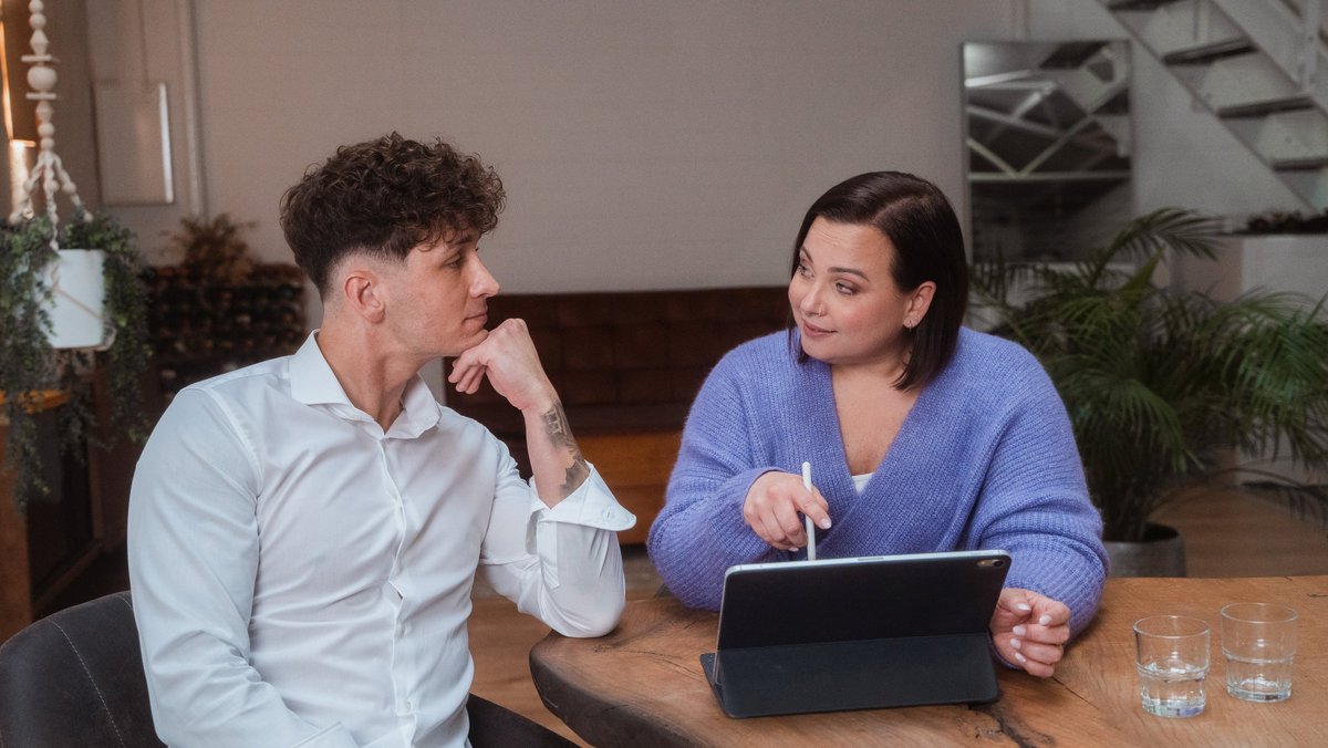 Un jeune homme et une jeune femme sont assis ensemble à une table. Devant eux se trouve une tablette, et ils se regardent avec confiance.