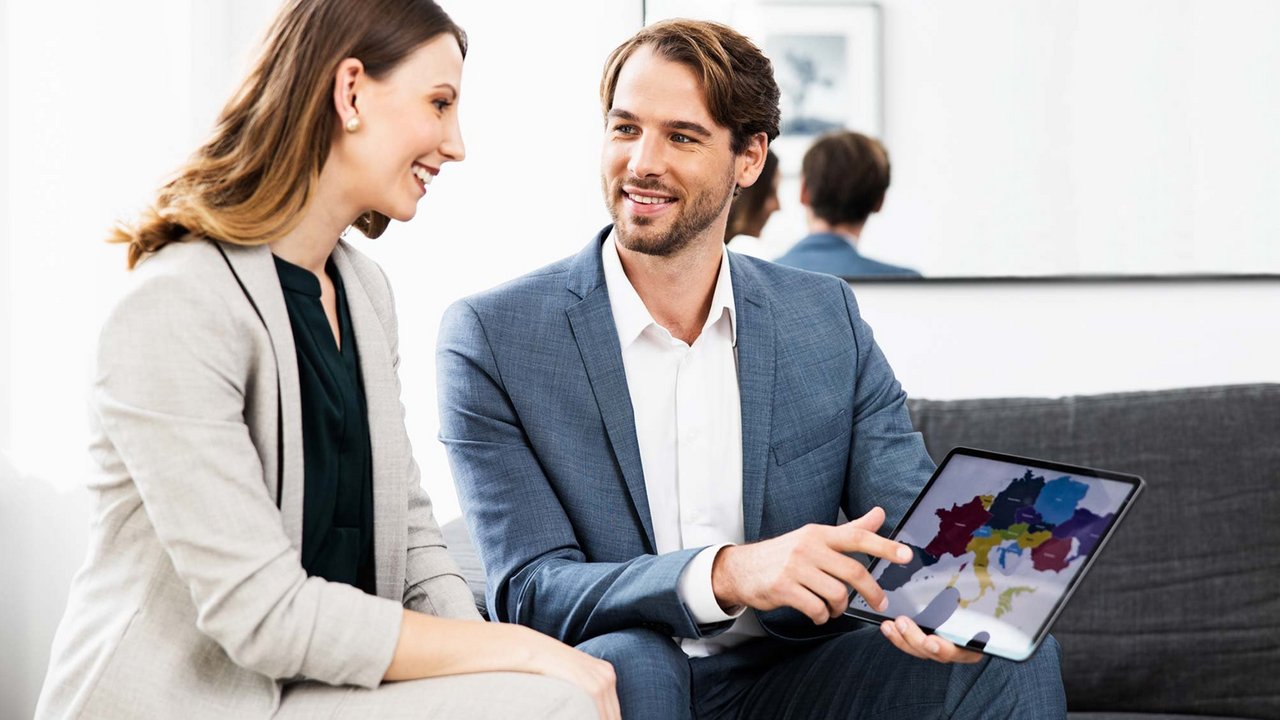 Un homme et une femme en tenue professionnelle sont assis sur un canapé et regardent ensemble une carte colorée de l'Europe sur une tablette.