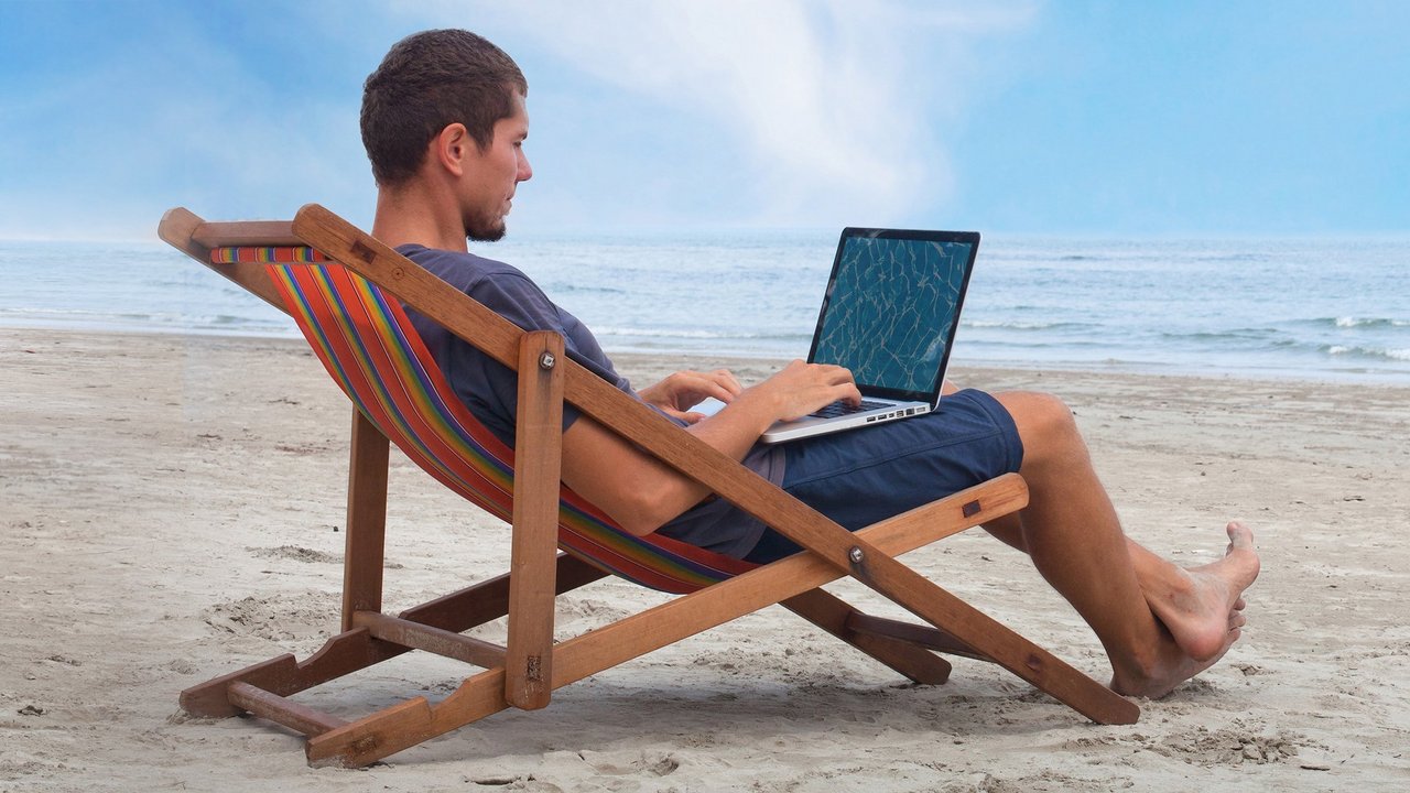 Un homme est assis sur une chaise longue à la plage et fait des recherches sur son ordinateur portable. 