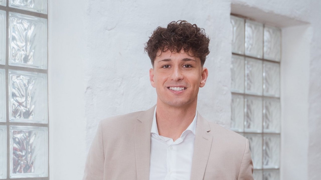 Un jeune homme en costume gris sourit aimablement à la caméra.