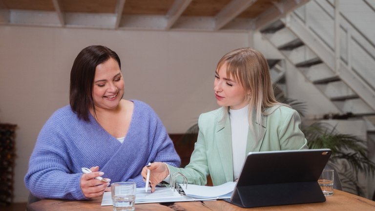 La femme de gauche semble satisfaite et reçoit des conseils de la femme à droite, qui est assise devant un ordinateur portable. Toutes deux regardent ensemble un document.